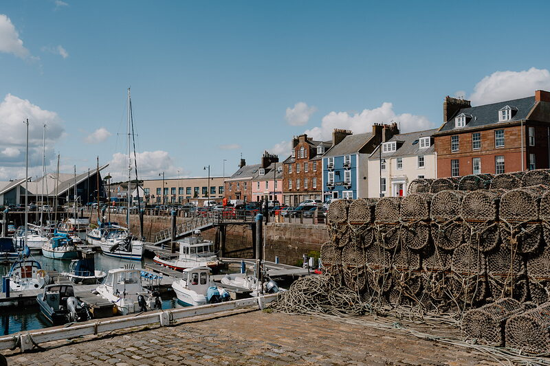 Boats and yachts fill a harbour beside stacked lobster creels, colourful terraced houses lining the quay behind.