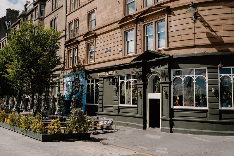 A green-fronted corner pub with arched windows stands beneath a tenement building, tables and chairs set out on the pavement beside young trees.