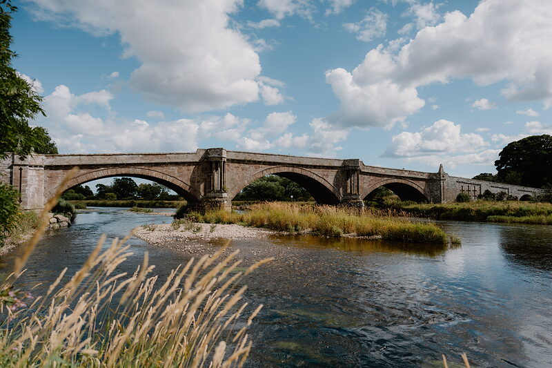 A stone road bridge with three arches crosses a shallow river lined with reeds and gravel banks under a blue sky.