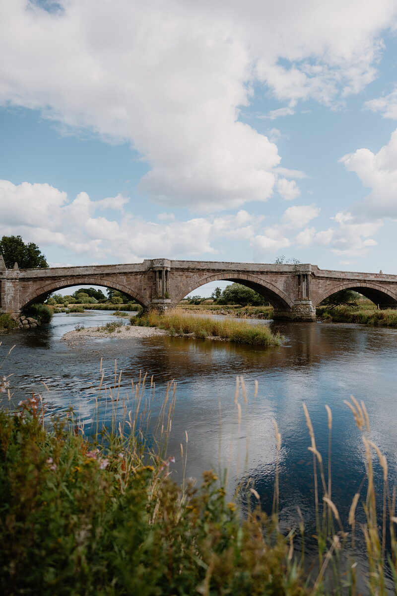A stone road bridge with four arches crosses a wide river lined with reeds and gravel islands under a partly cloudy sky.