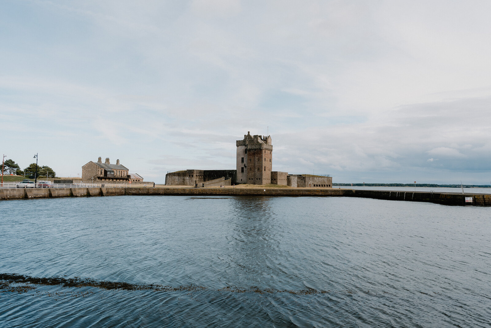 A stone tower with battlements stands on a small tidal promontory between two harbour walls, calm water reflecting the sky around it.