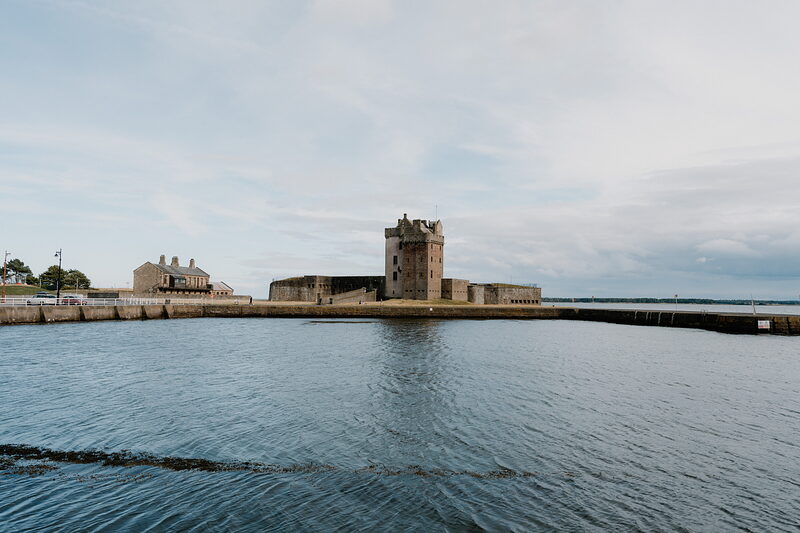A stone tower with battlements stands on a small tidal promontory between two harbour walls, calm water reflecting the sky around it.