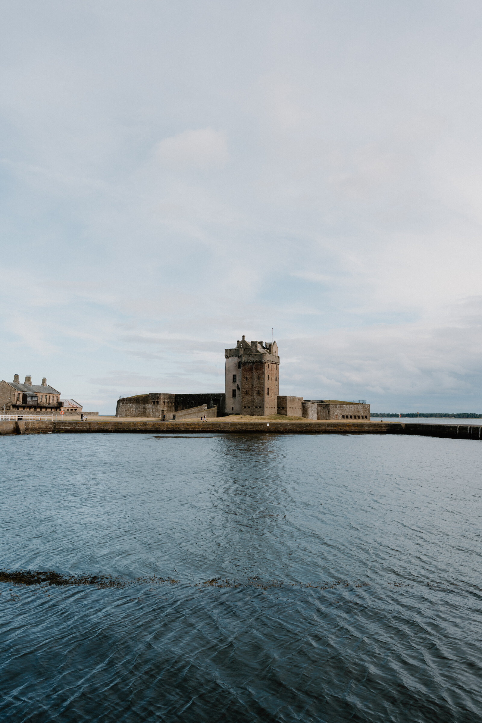 A stone tower with battlements stands on a small tidal promontory between two harbour walls, calm water and pale evening sky around it.