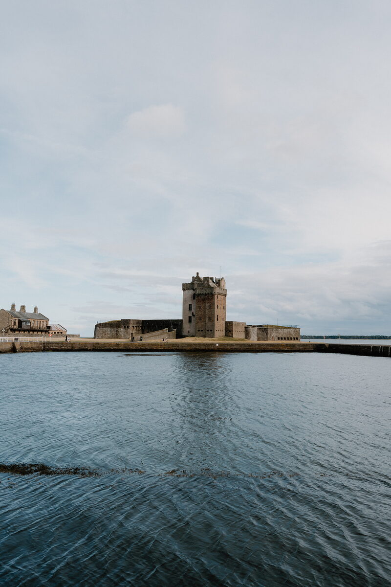 A stone tower with battlements stands on a small tidal promontory between two harbour walls, calm water and pale evening sky around it.