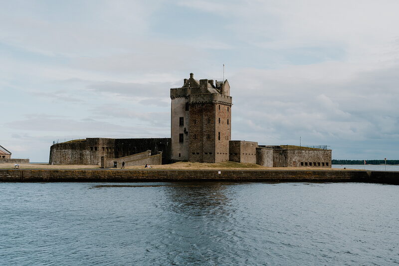 A stone tower with battlements stands on a small tidal promontory between two harbour walls, calm water reflecting the overcast sky.