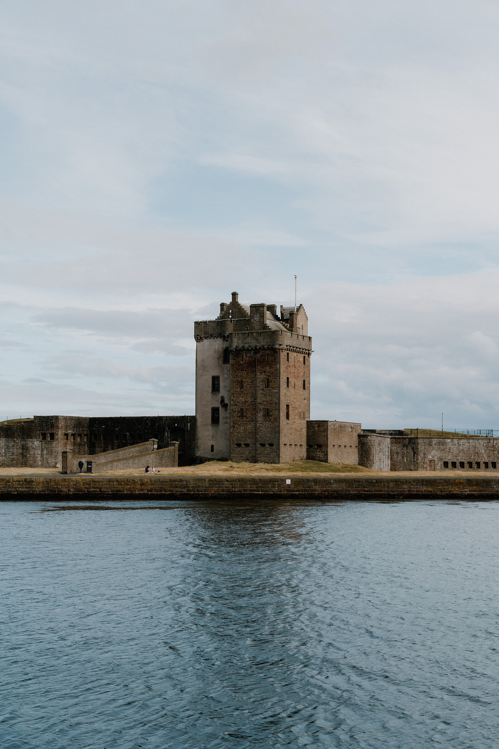 A stone tower with battlements stands on a small tidal promontory between two harbour walls, calm water reflecting the sky and a person visible on the shore path.
