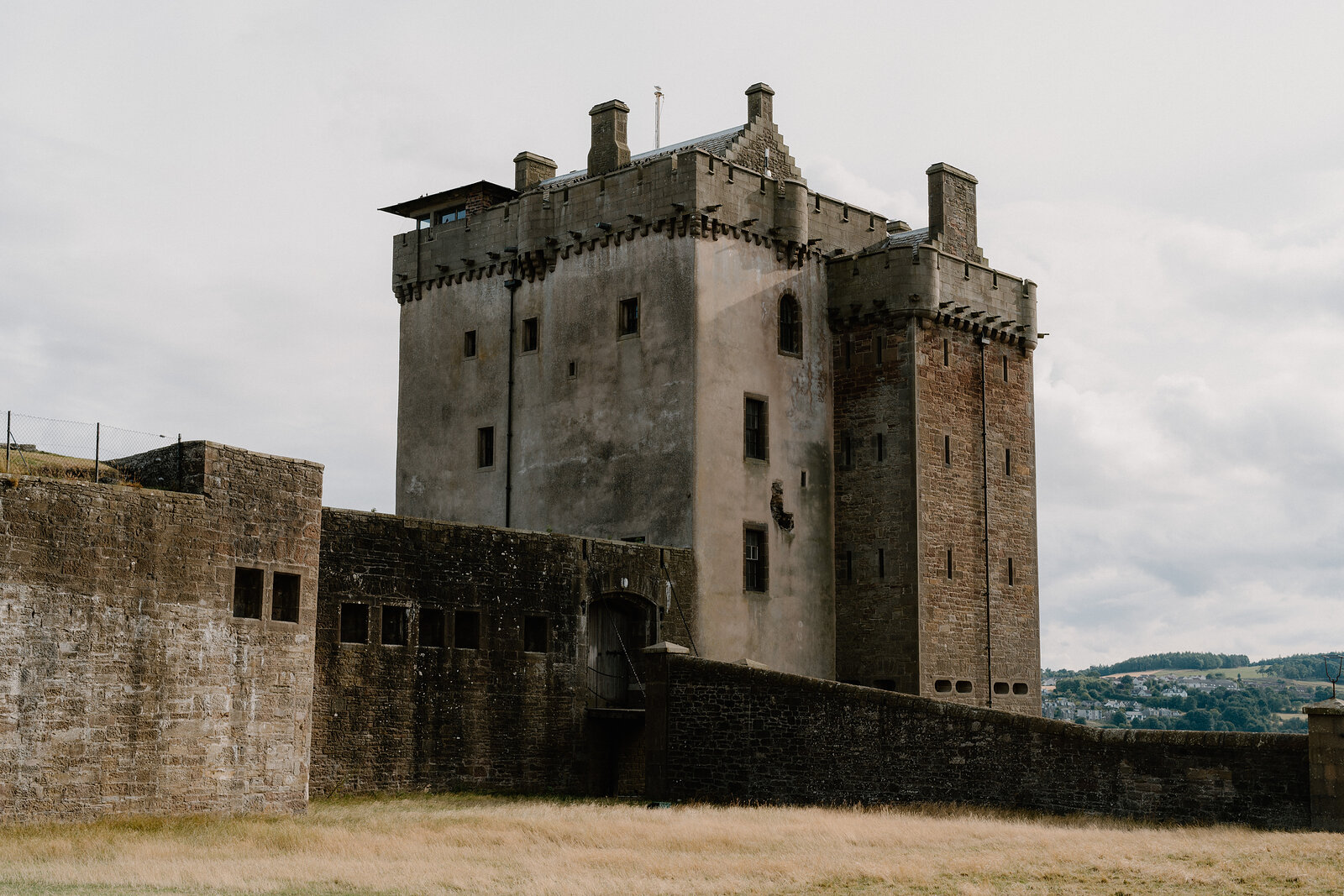 A rendered stone tower castle with battlements and a projecting stair turret rises behind a fortified curtain wall.