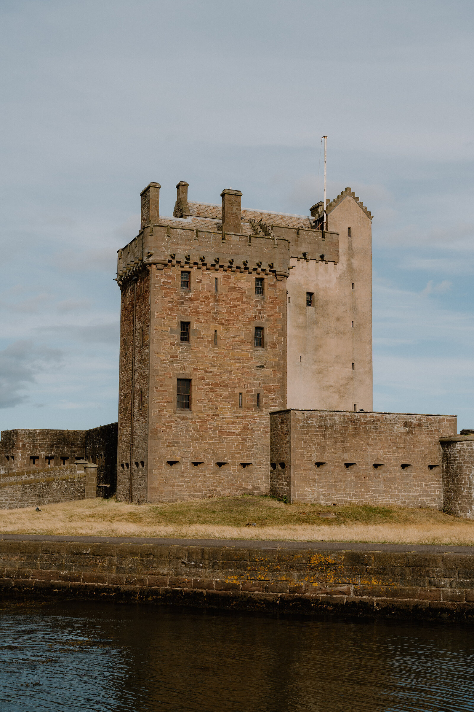 A rendered stone tower castle with battlements and a flagpole rises behind a fortified curtain wall beside water.