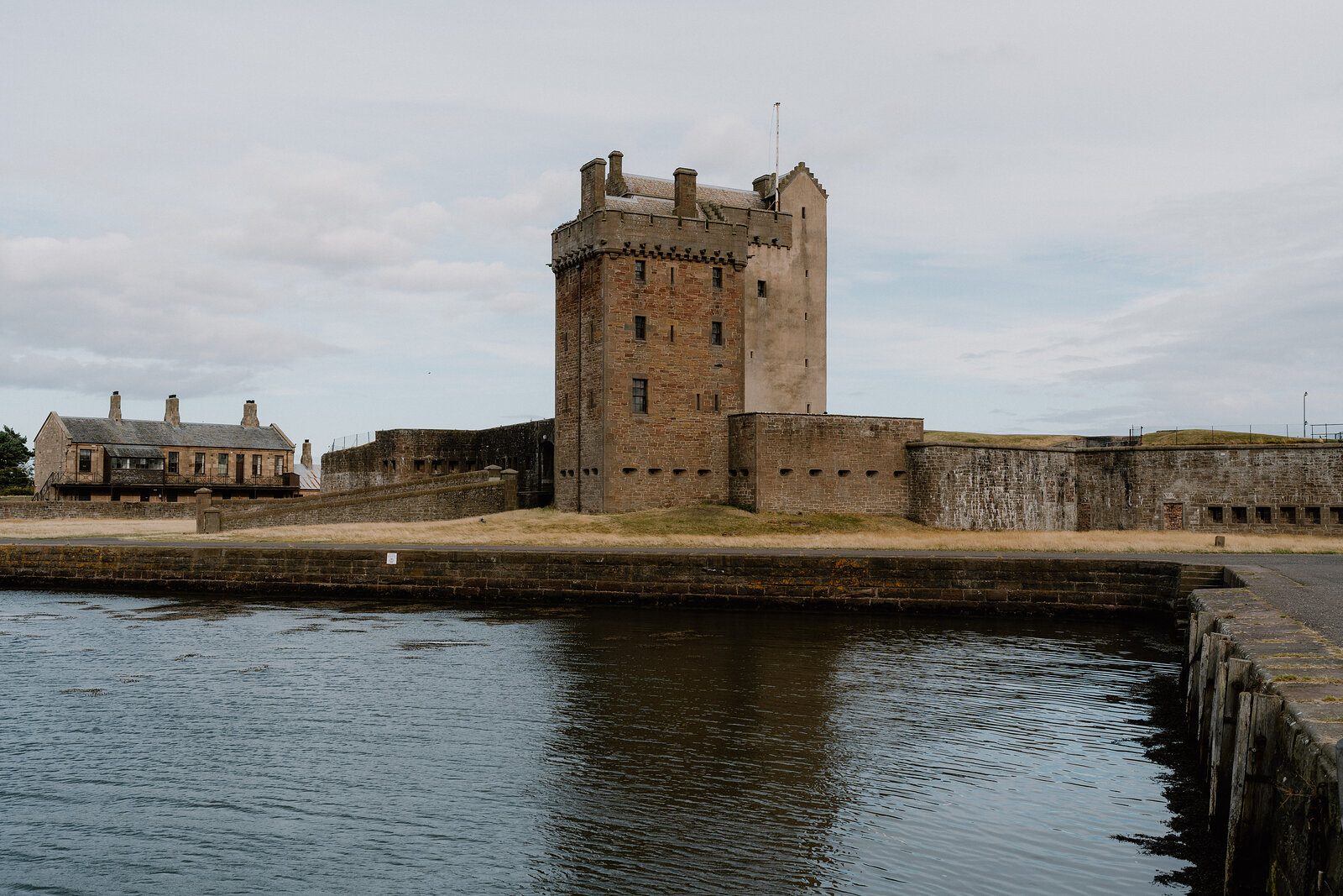 A rendered stone tower castle with battlements rises behind a fortified curtain wall, a low building standing beside the water in front.