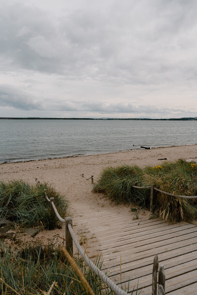 A wooden boardwalk with rope railings leads through dune grass onto a wide sandy beach beside calm sea.