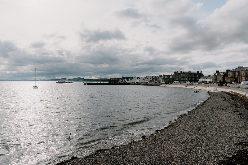A pebble beach curves along calm water toward a distant pier and town, a moored sailing boat and clouds filling the sky.