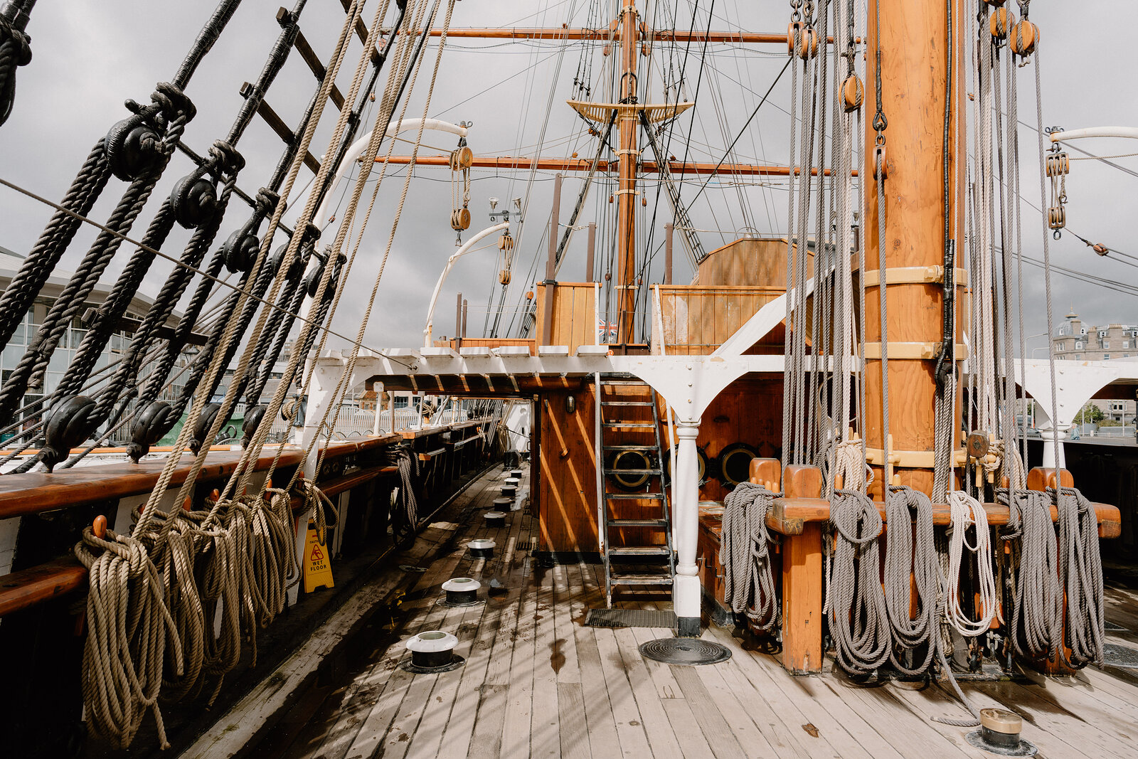 The deck of a tall ship shows a raised walkway, ladder and coiled ropes hanging from wooden rails, masts and rigging rising above.