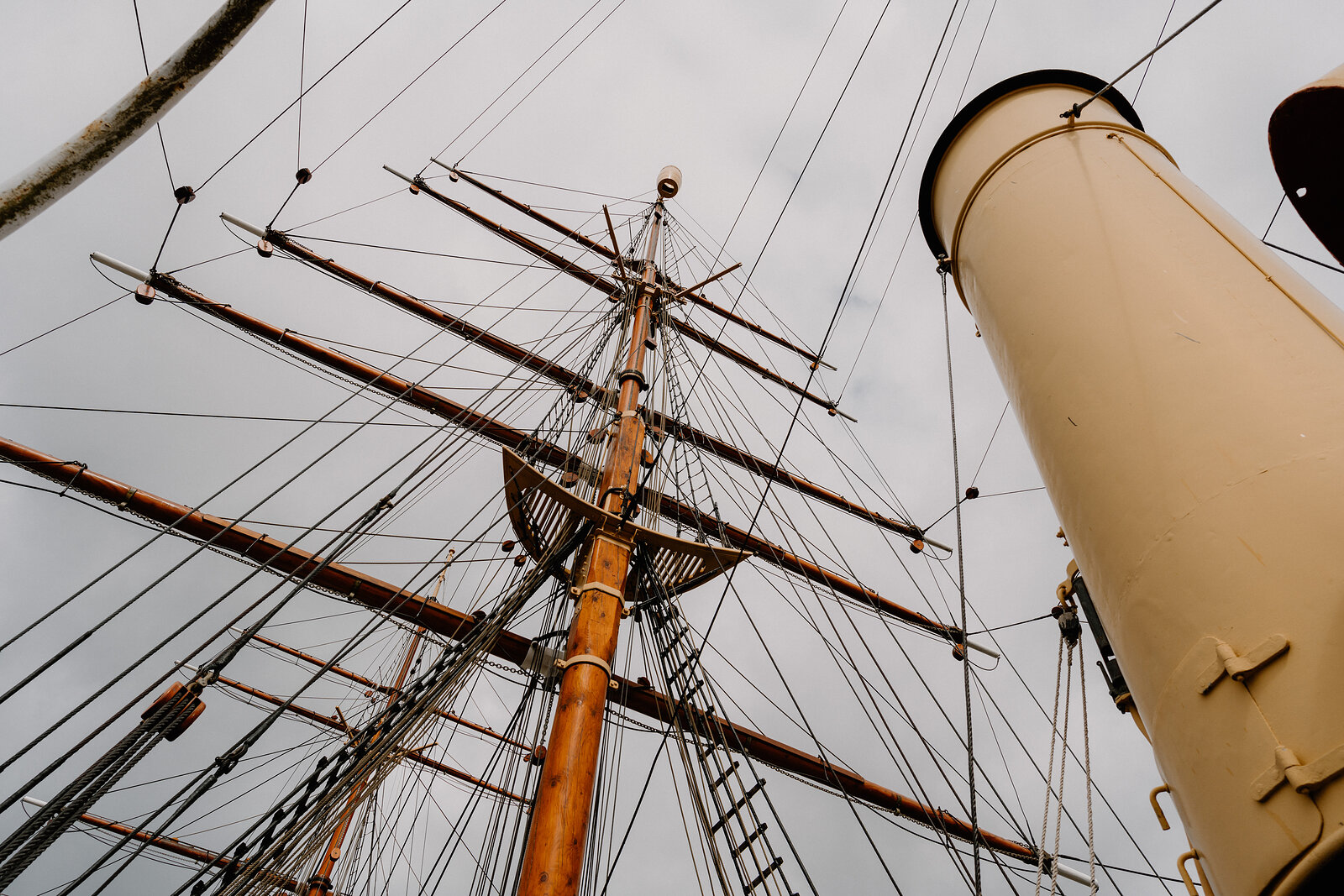 Looking up at a ship's mast and rigging beside a tall cream funnel against an overcast sky.