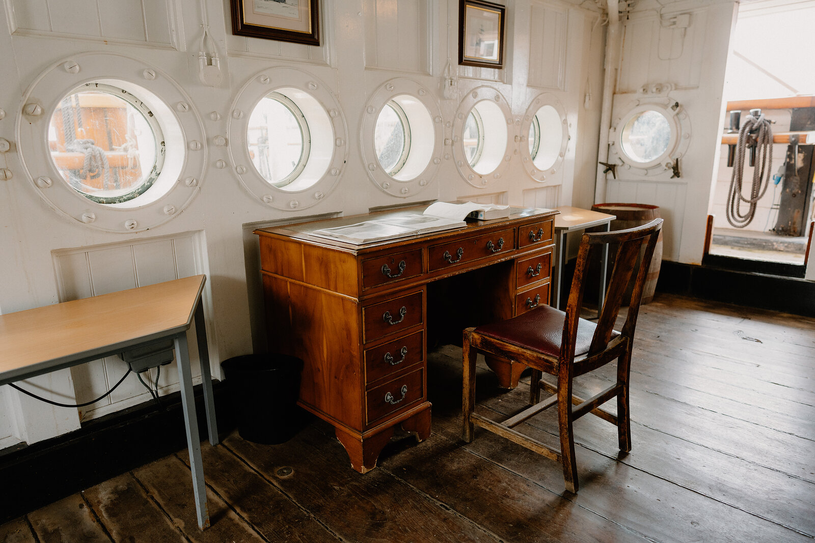 An antique writing desk and chair stand beneath round brass portholes in a panelled ship cabin, a small table to one side.