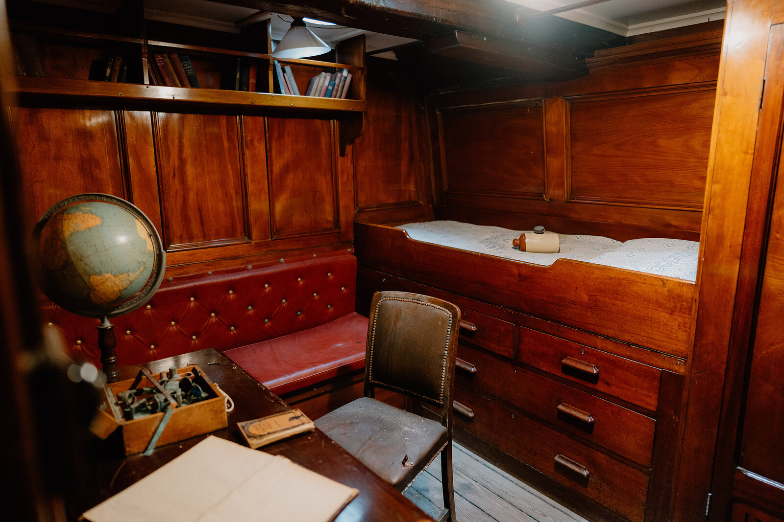 A built-in bunk with drawers stands beside a red leather bench, a globe and writing desk furnishing a wood-panelled ship cabin.