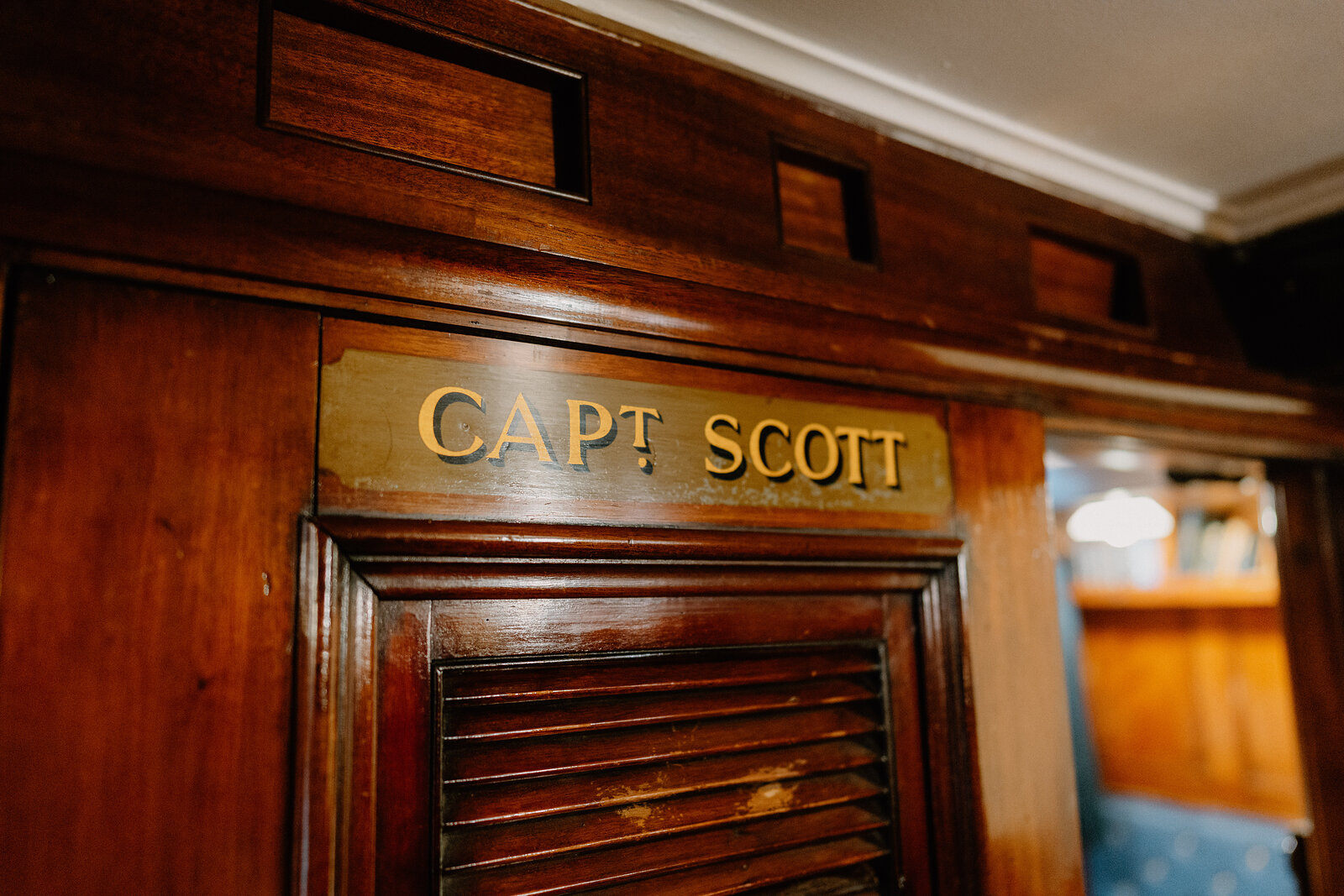 A brass nameplate reading "Capt Scott" is mounted above a louvred wooden door in a ship's panelled corridor.