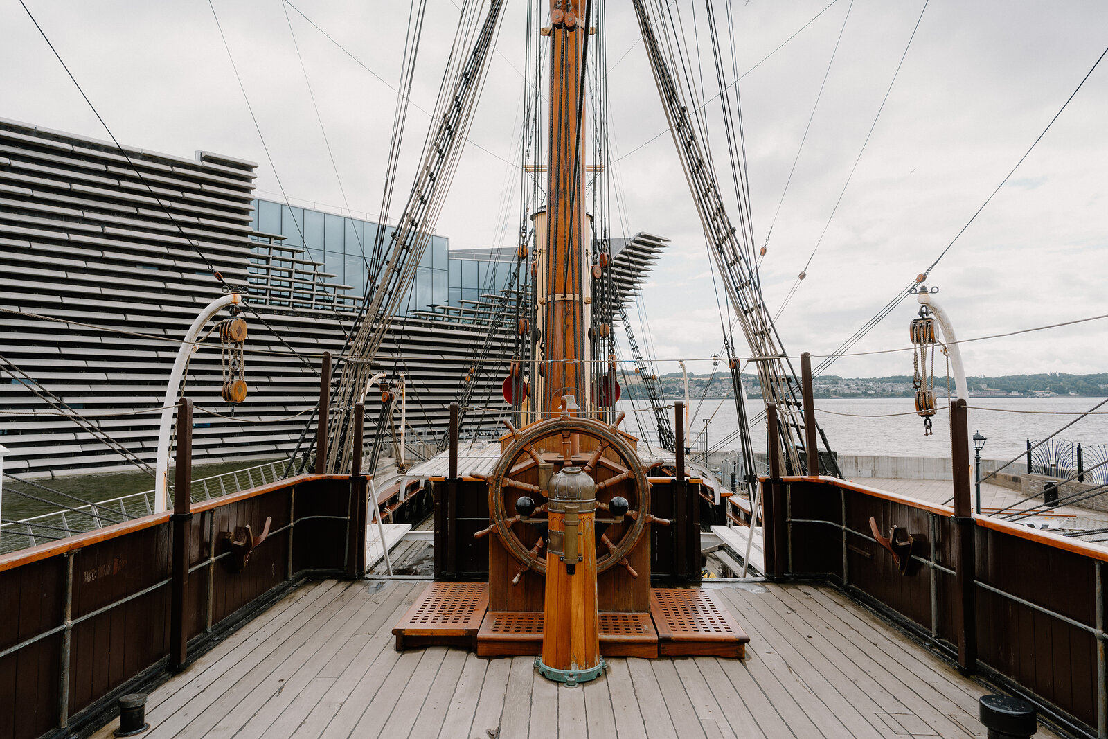 A ship's wooden wheel and mast rise from the deck, rigging stretching overhead and a modern glass and steel building visible on the quayside behind.