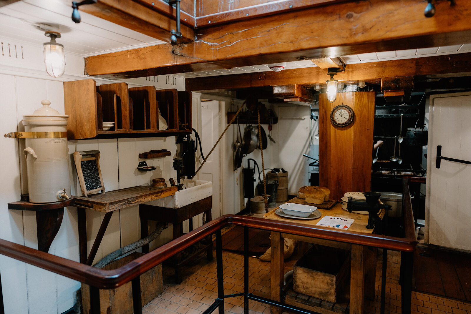 A ship's galley holds a wooden counter, milk churn, wall clock and metal pipework beneath exposed beams, a dark stove visible at the far end.