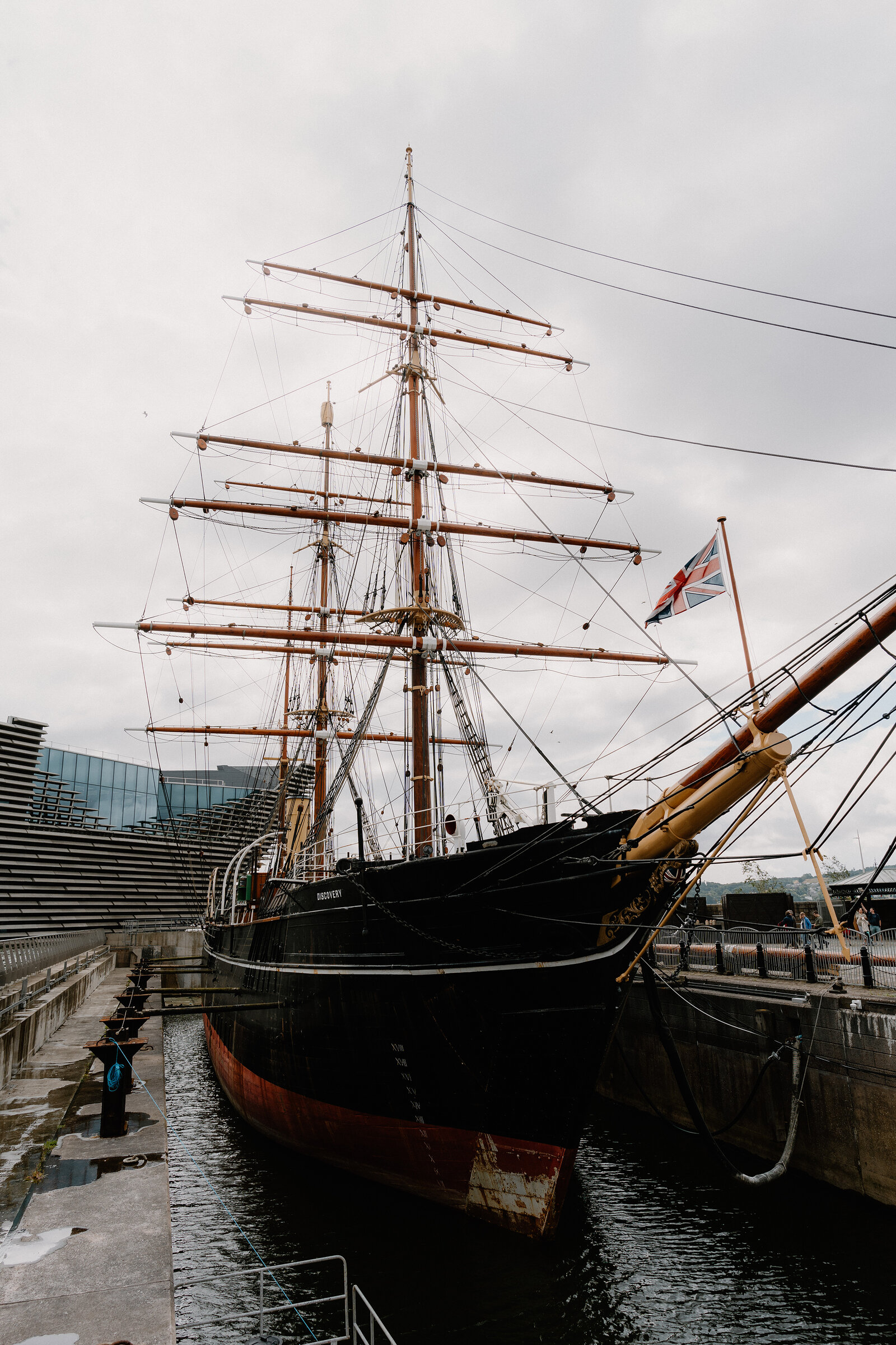A tall ship with rigged masts and a Union flag sits in dry dock beside a modern glass and steel building, a person walking along the quayside.