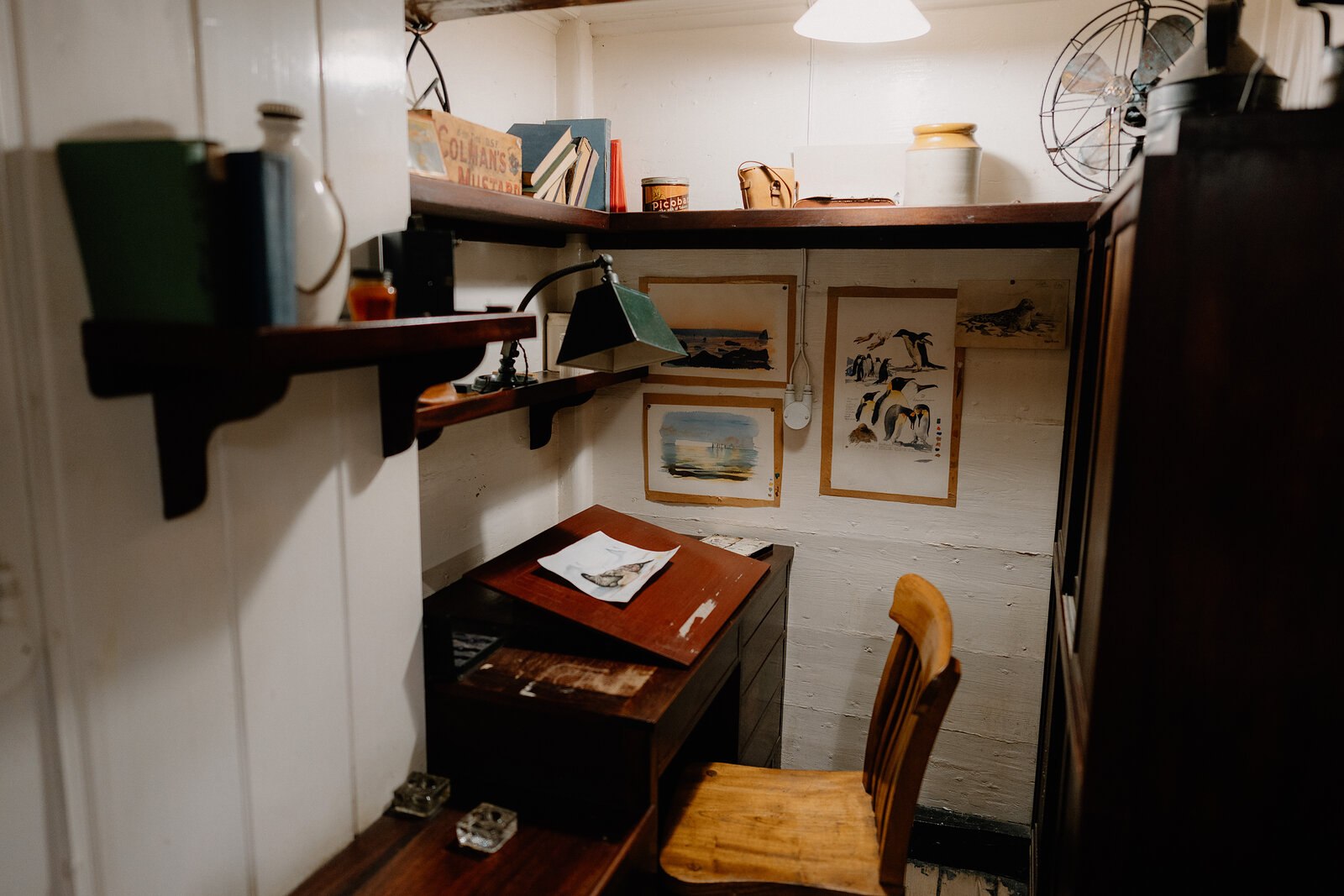 A small wood-panelled study nook holds a writing desk, chair and shelving of books, framed prints of ships and landscapes hanging on the wall.