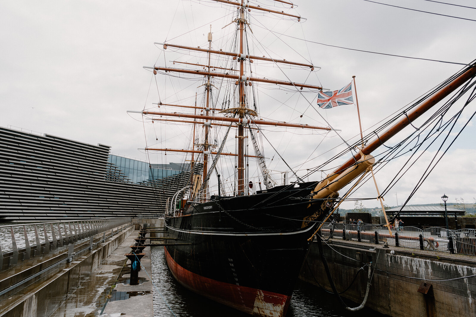 A tall ship with rigged masts and a Union flag sits in dry dock beside a modern glass and steel building.