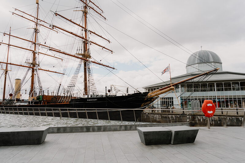 A tall ship with rigged masts sits in dry dock beside a domed building, a red life ring mounted on a stone bench in the foreground.