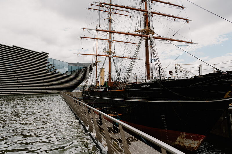 A tall ship with rigged masts sits in dry dock beside a wave-patterned modern building, a wooden walkway with railings leading toward it.