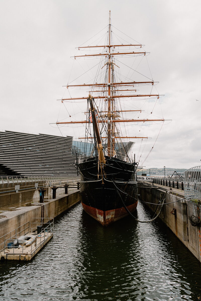 A tall ship's bow rises in dry dock, rigged masts stretching overhead beside a wave-patterned modern building.