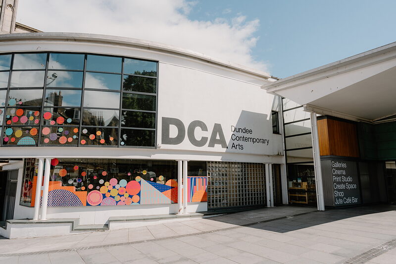 A building lettered "DCA, Dundee Contemporary Arts" displays a colourful abstract mural beside its glazed entrance, a sign listing galleries, cinema and cafe bar.
