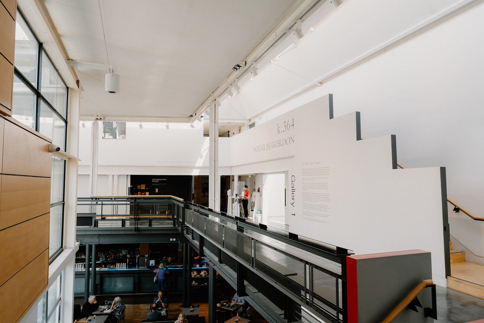 A white gallery interior with an angled staircase and mezzanine overlooks a lower floor, a sign reading "Gallery" mounted on the wall and visitors walking below.