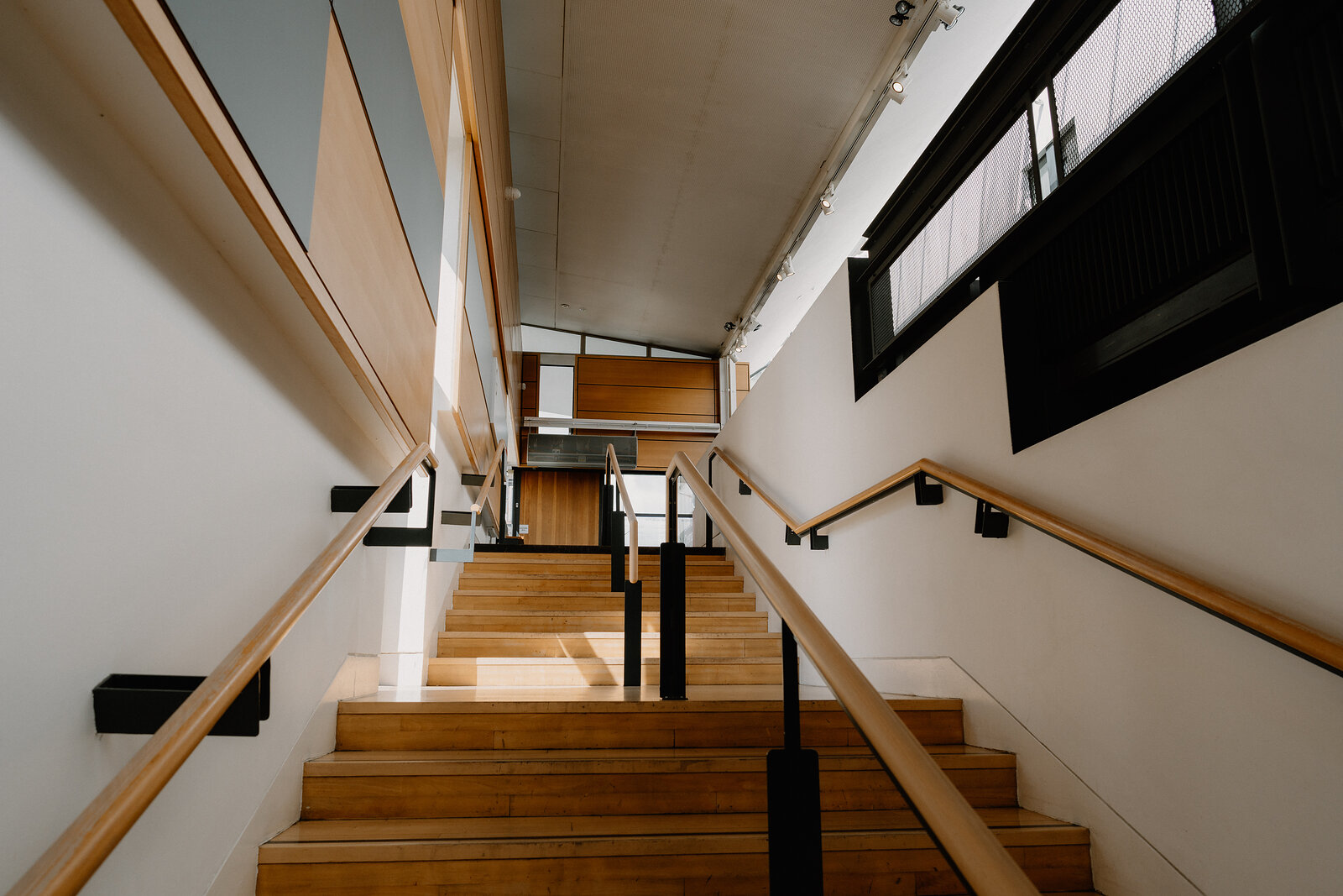 A wooden staircase descends between glass-panelled walls in a gallery interior, handrails lining both sides.