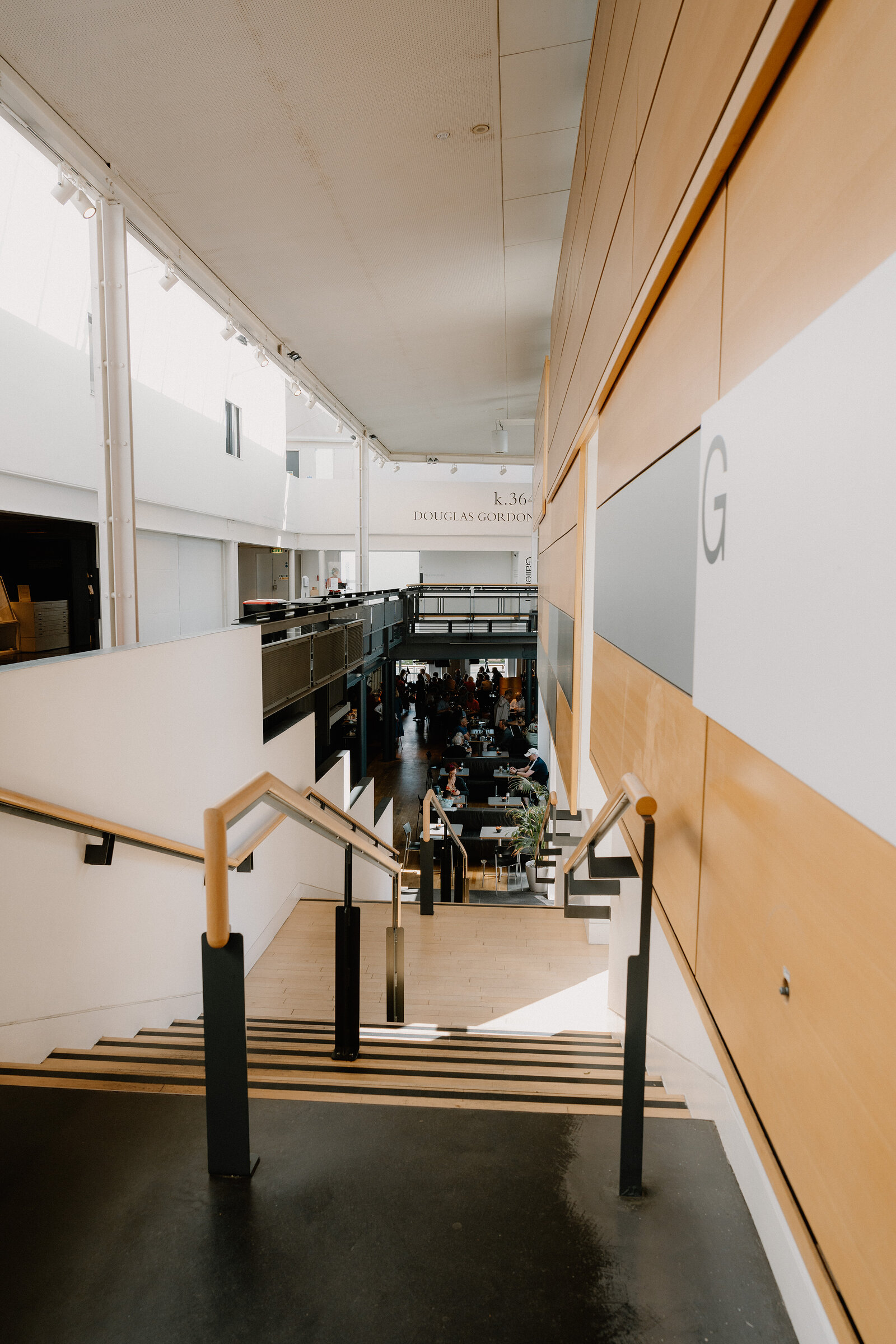 A wooden staircase descends past a sign reading "Douglas Gordon" toward a busy café below, visitors seated at tables.