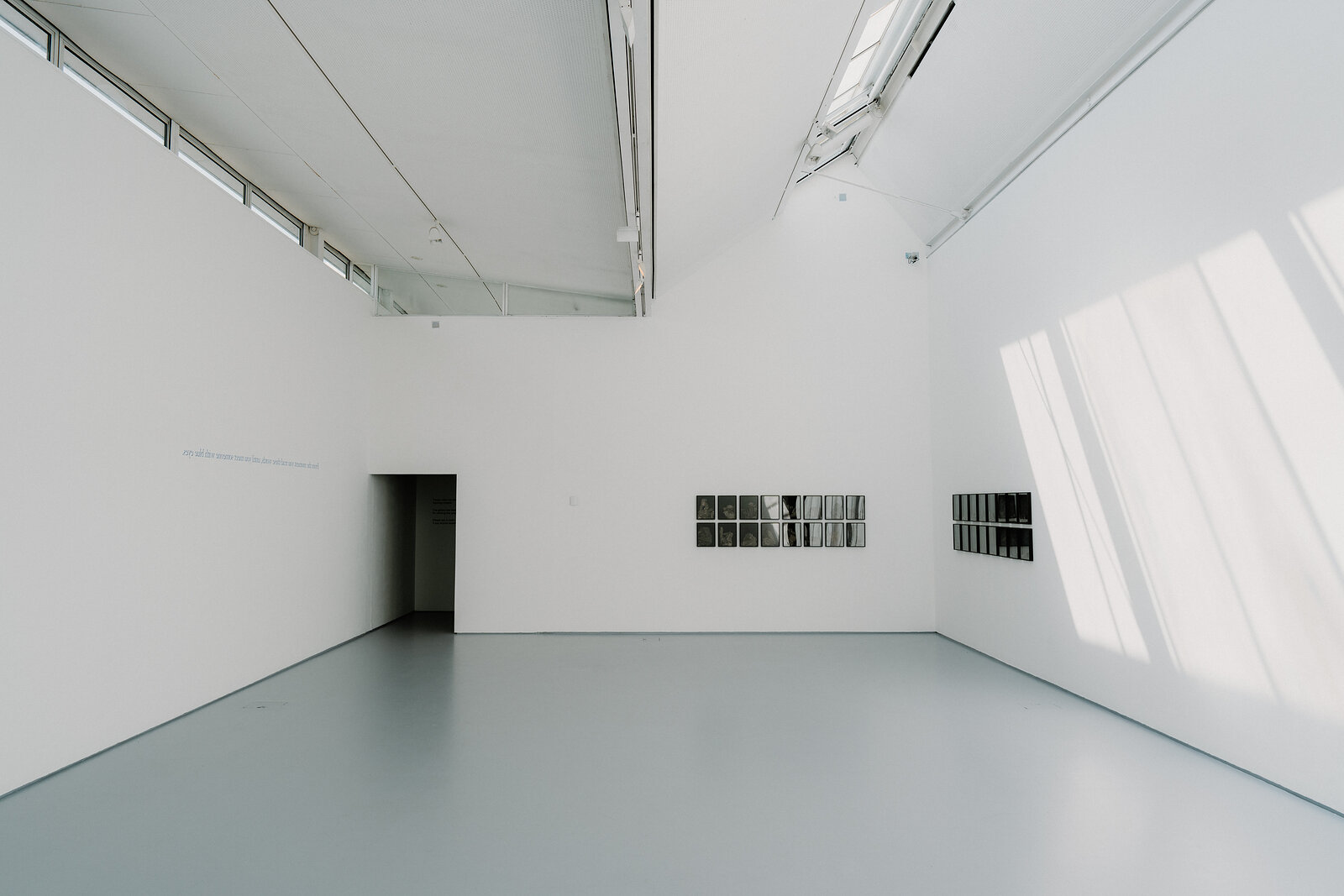 A pale gallery room holds two rows of small framed black and white prints on adjoining white walls.