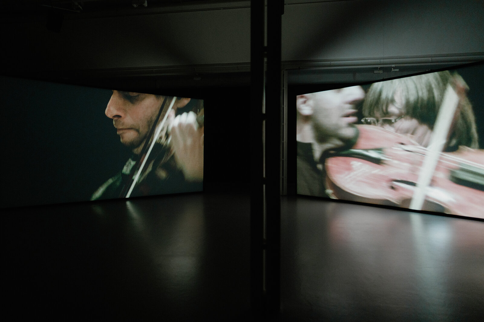 Two large video screens show close-up footage of musicians playing violins in a dark gallery installation.