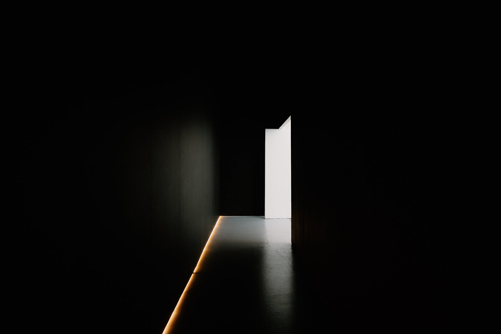 A single open door glows with light at the end of a completely dark corridor.