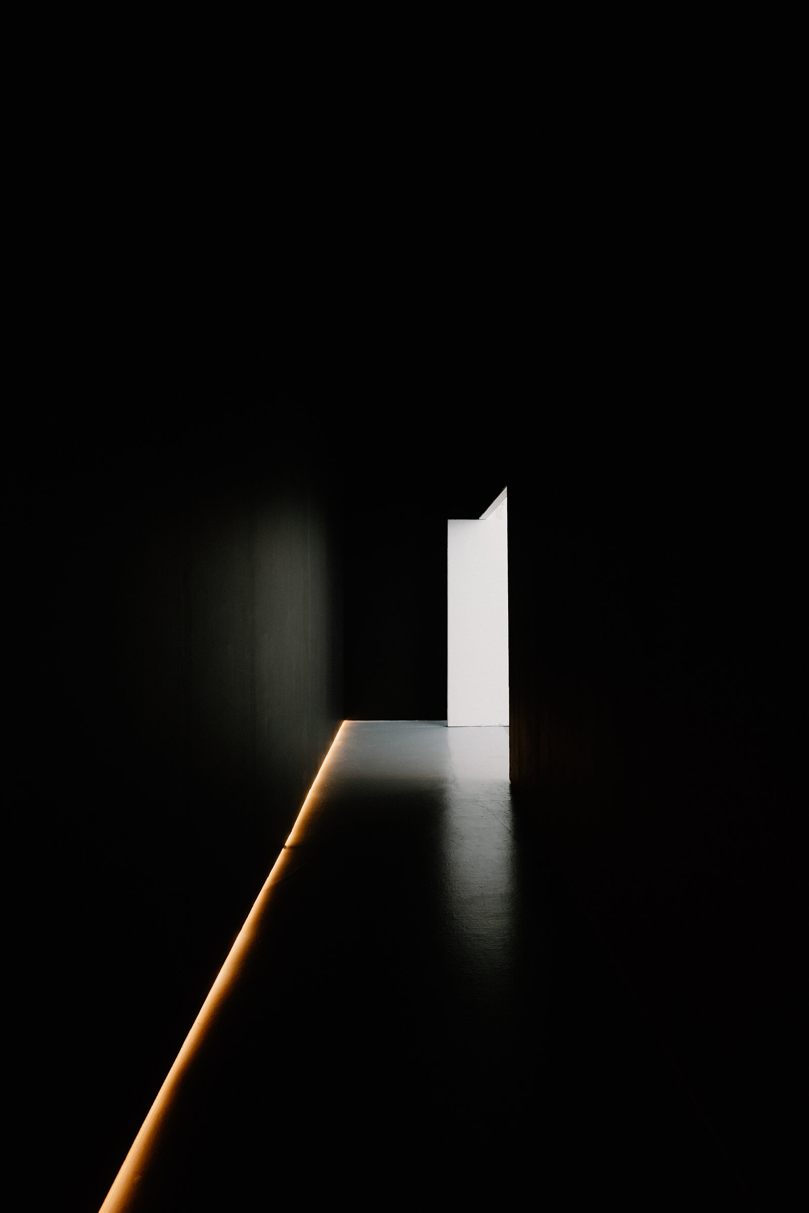 A single open door glows with light at the end of a completely dark corridor.