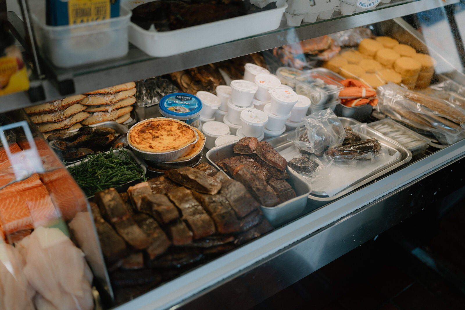 A refrigerated counter displays smoked fish fillets, a quiche, wrapped cheeses, dips and pâté, with foil-topped tubs and packaged pastries stacked above.