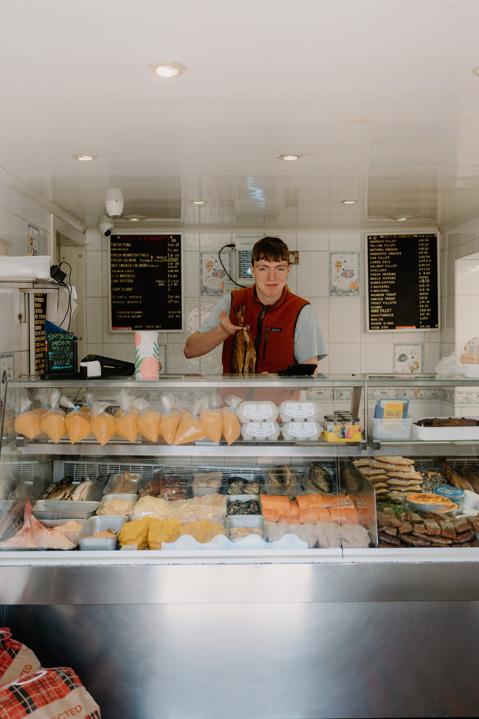 A young man in a red waistcoat holds up a whole fish behind a counter stocked with prawns, eggs, sandwiches and packaged fish, price boards listing items on the wall behind him.