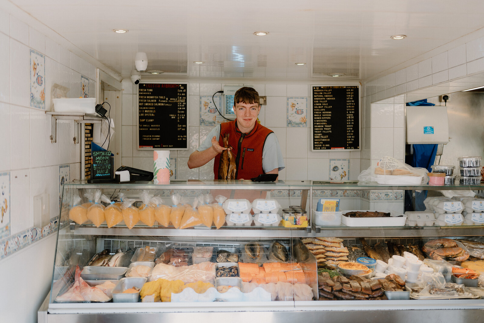 A young man behind a fishmonger's counter holds up a whole fish, trays of prawns and boiled eggs and menu boards visible around him.
