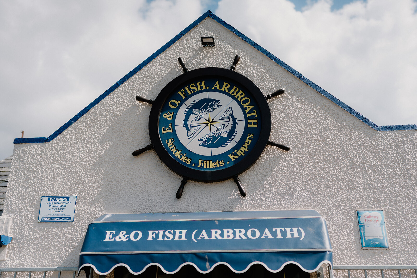 A ship's wheel sign reading "E & O FISH ARBROATH — Smokies, Fillets, Kippers" is mounted on a white pebble-dashed gable above a green awning that reads "E&O FISH (ARBROATH)".