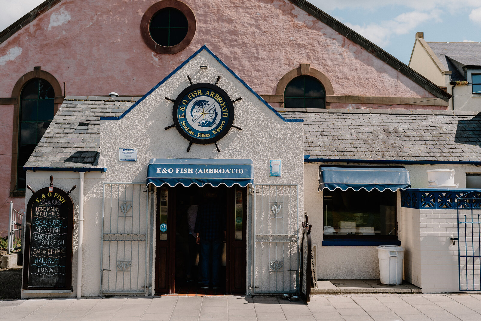 The pink gabled front of "E&O FISH (ARBROATH)" has a ship's wheel sign above the doorway, a chalkboard menu and blue awning beside the barred entrance.