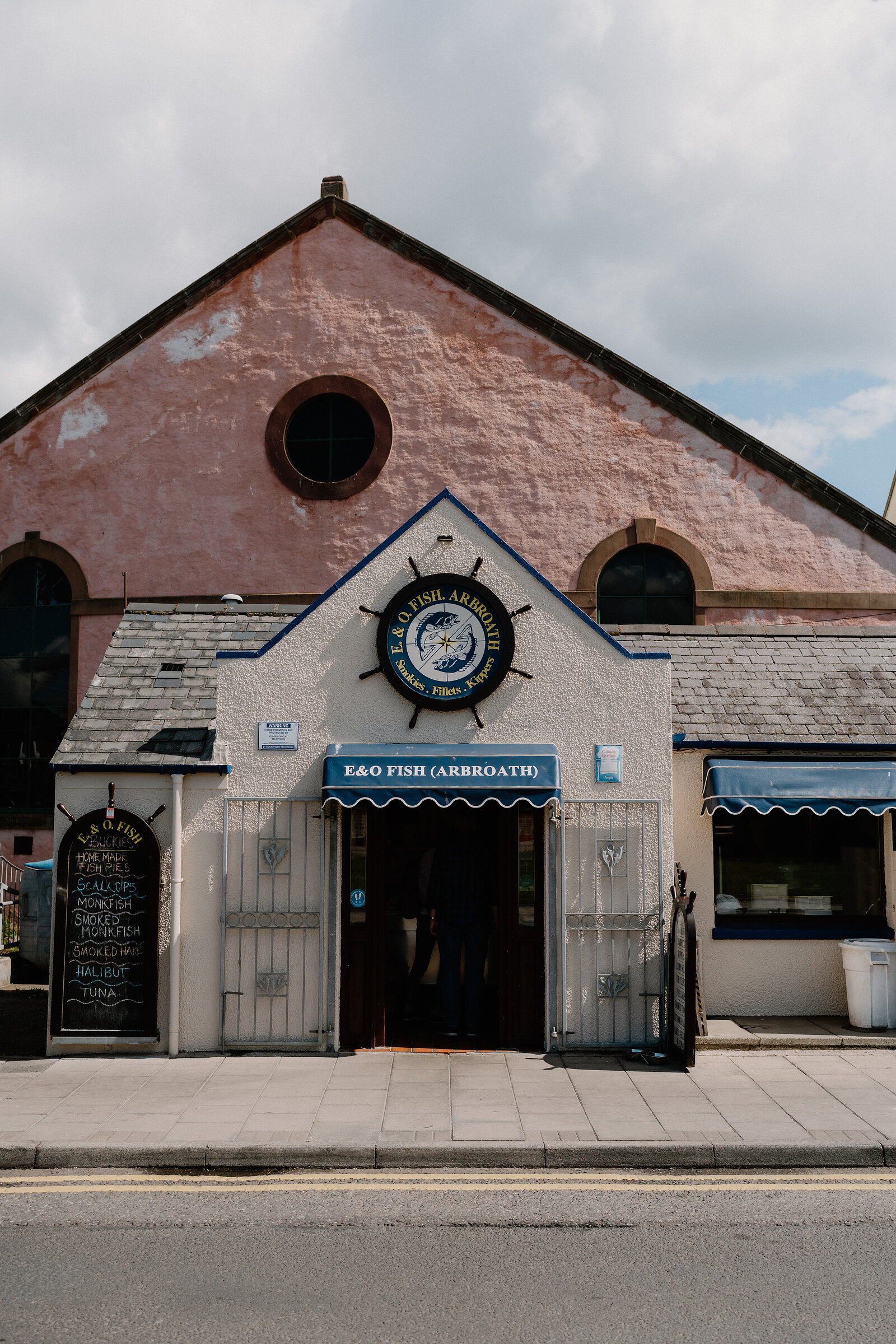 The pink gable of "E&O FISH (ARBROATH)" carries a ship's wheel sign and a round window, a blue awning and barred doorway beneath a chalkboard menu.