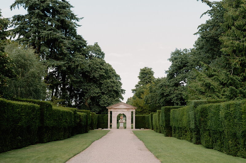 A gravel path runs between tall clipped yew hedges towards a small classical stone pavilion at the far end of a formal garden.