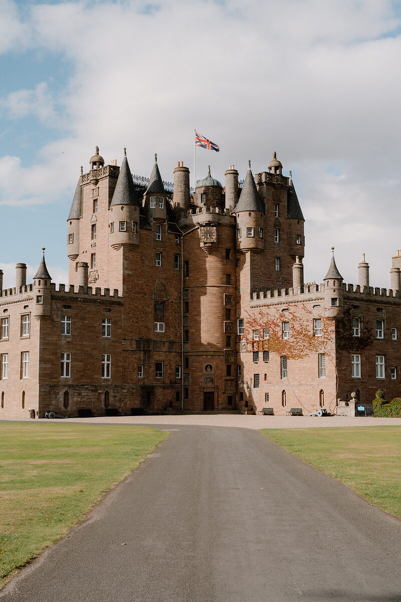 A turreted red sandstone castle flies a flag above its ornate skyline, a gravel driveway leading up to the entrance between mown lawns.
