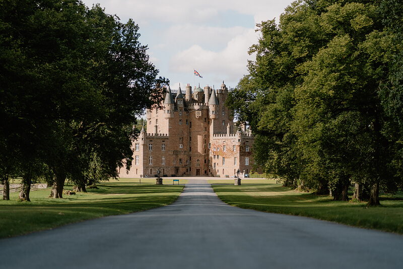 A turreted red sandstone castle stands at the end of a tree-lined avenue, mown lawns stretching on either side.