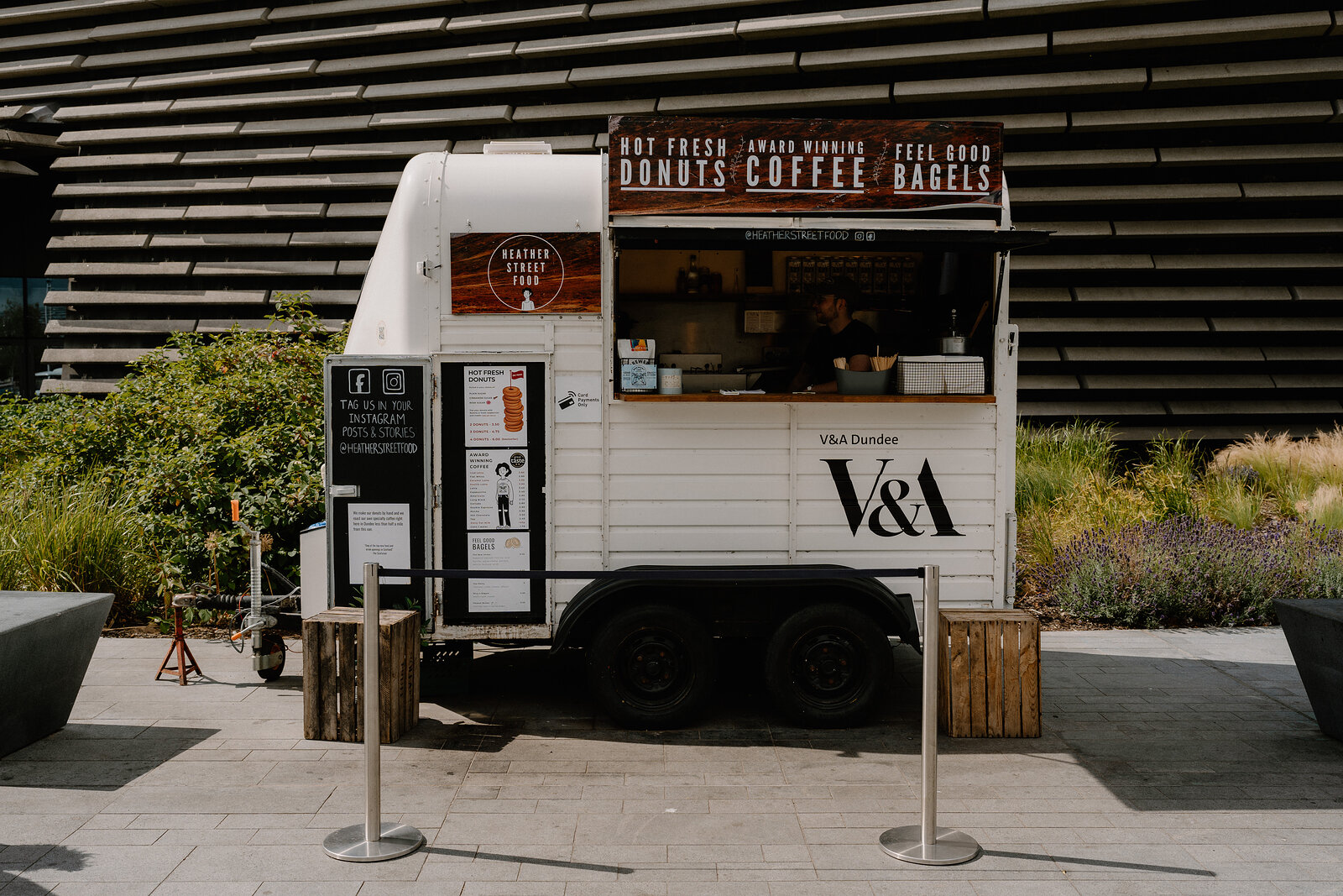 A converted horsebox food truck branded "V&A Dundee" advertises "HOT FRESH DONUTS", "COFFEE" and "BAGELS" on its serving hatch, potted plants standing either side.