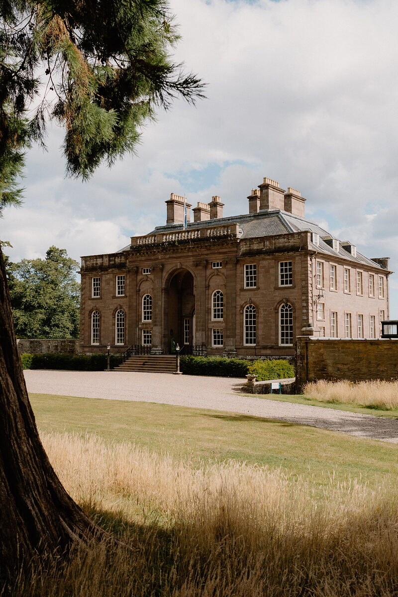A grand sandstone mansion with a pedimented entrance stands beyond a gravel drive, a mature cedar tree framing the view.