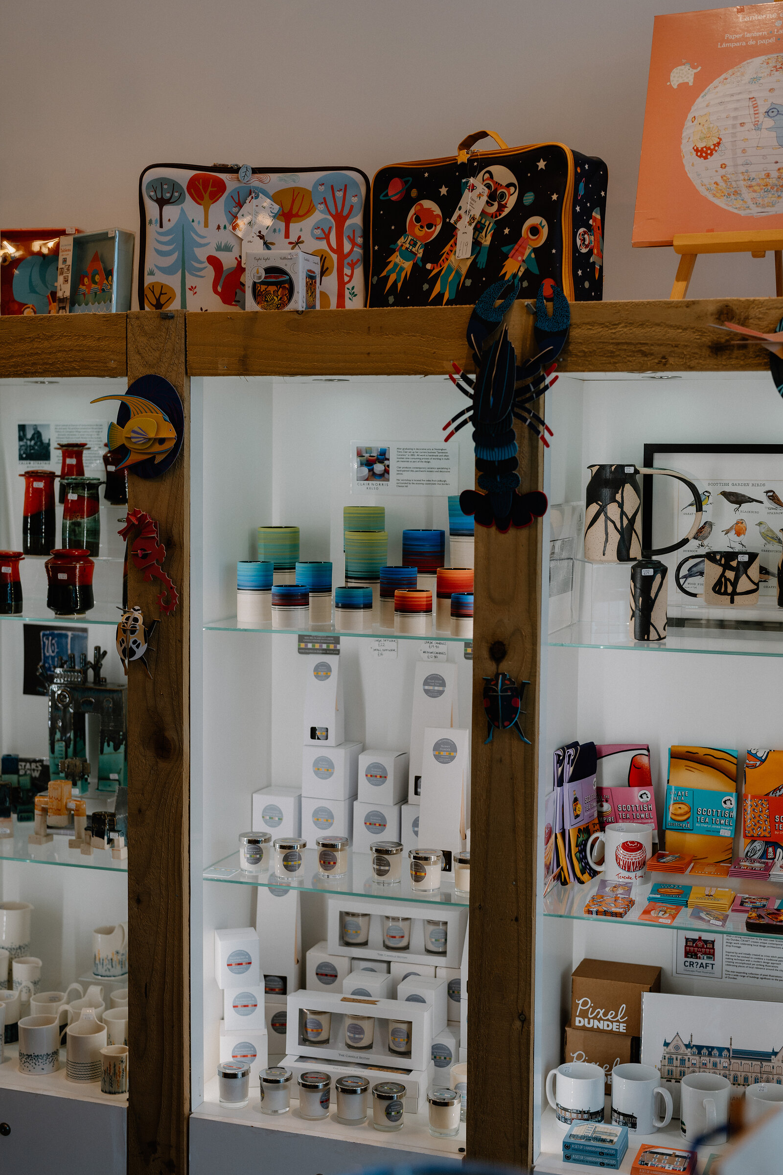 Glass shelving displays candles, mugs and greeting cards in a gift shop, a mug printed "Pixel Dundee" standing among framed prints and colourful bags.