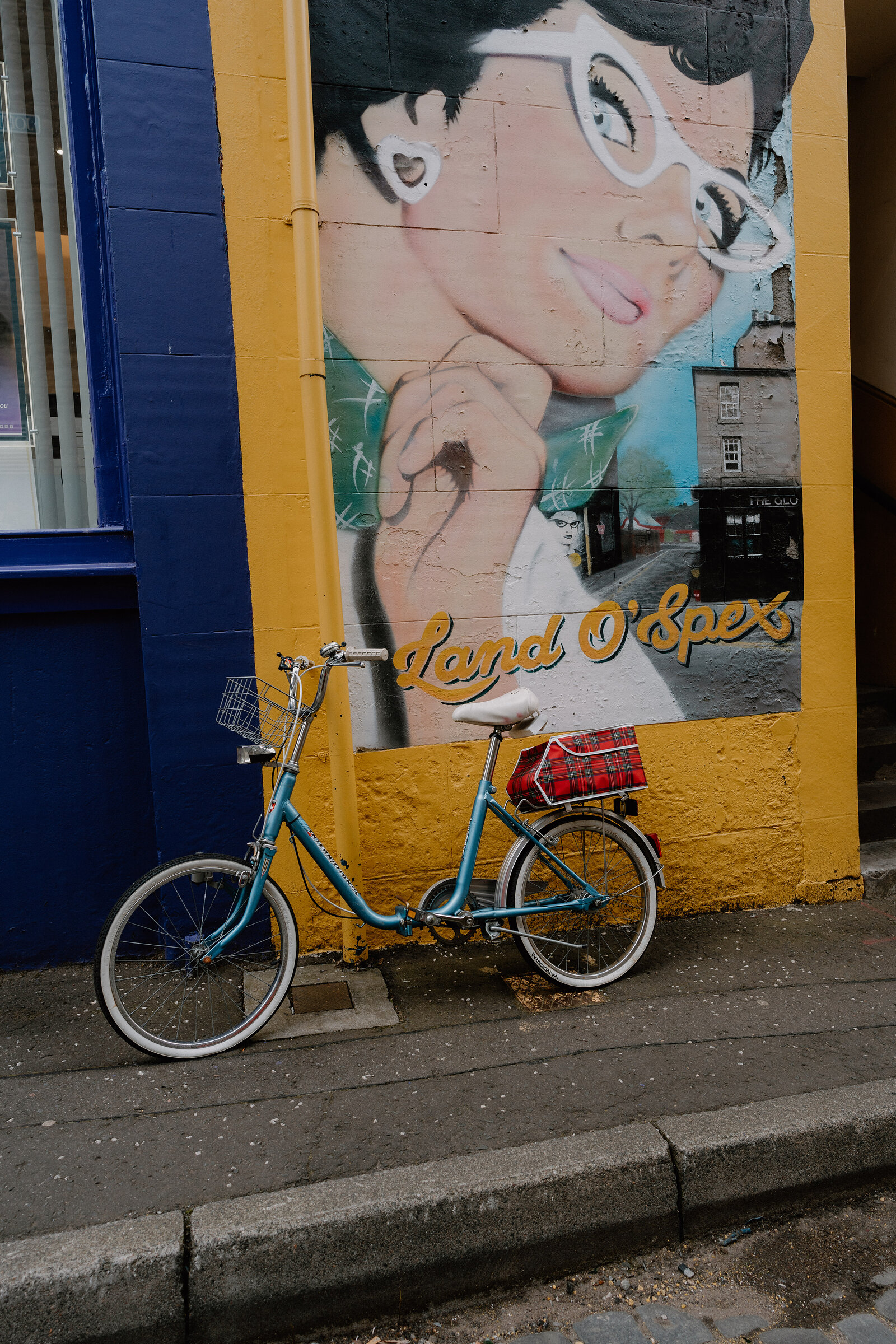 A mural titled "Land O'Spex" depicts a woman in cat-eye glasses beside a small townscape, a vintage bicycle parked in front of the painted wall.