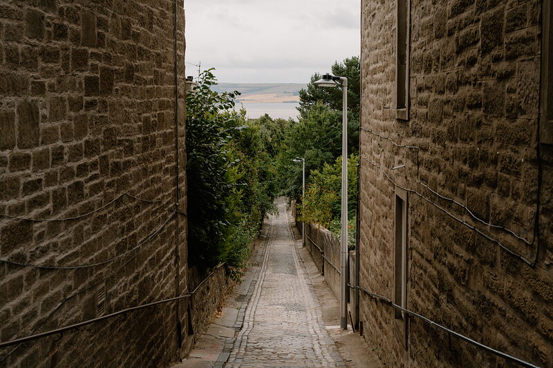 A narrow cobbled lane descends steeply between stone buildings towards a distant firth and hills.